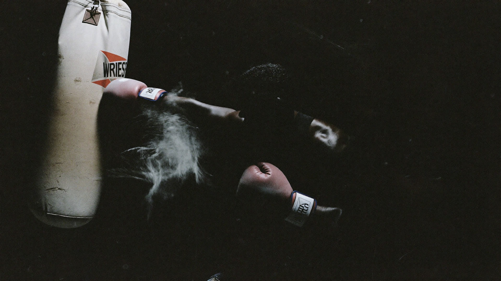 Boxer mid-punch on a heavy bag under harsh gym light, chalk dust in the air